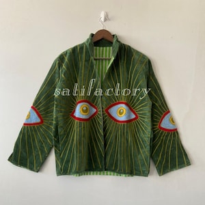 May include: Green velvet jacket with embroidered eye designs and radiating lines. The jacket has a high collar, long sleeves, and a striped green and white lining. The text "salifactory" is visible on the jacket.