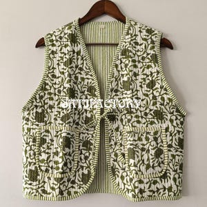 Può includere: Un gilet trapuntato senza maniche con un motivo floreale nei toni del verde e del bianco. Il gilet ha due tasche anteriori e una fodera a righe verdi e bianche. È appeso a una gruccia di legno.