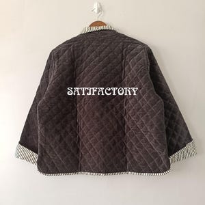 以下が含まれることがあります： ダイヤモンド模様のダークブラウンのキルティングジャケット。明るいグレーの背景に掛けられています。襟と袖口にはストライプのトリムが付いています。「SATISFACTORY」の文字がジャケットの背面にプリントされています。