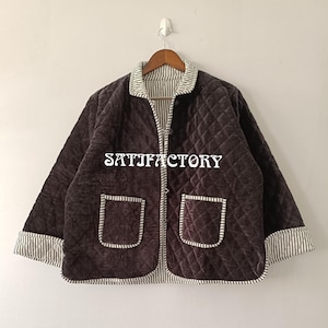 以下が含まれることがあります： 茶色のキルティングジャケット。ダイヤモンド模様、2つのフロントポケット、ストライプの襟と袖口が付いています。 ジャケットには「SATISFACTORY」の文字がプリントされています。