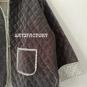 以下が含まれることがあります： ダイヤモンド模様のダークグレーのキルティングジャケット。 ジャケットには、白と黒のストライプ生地のポケットとトリムが付いています。 ジャケットには「SATISFACTORY」の文字がプリントされています。