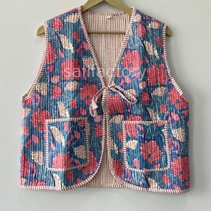 Puede incluir: Un chaleco acolchado con un estampado floral en tonos azules, rosas y blancos. El chaleco tiene dos bolsillos delanteros y un cierre de lazo. El forro interior presenta un patrón de rayas. El chaleco se muestra en una percha de madera.