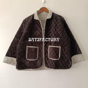 以下が含まれることがあります： ダイヤモンド柄のダークブラウンのキルティングジャケット。フロントポケットが2つ、ストライプの襟と袖口が付いています。ジャケットには「SATISFACTORY」の文字がプリントされています。