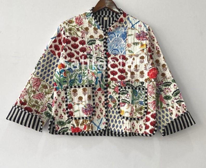 K&ouml;nnte beinhalten: Gesteppte Jacke mit Patchwork-Design mit floralen und geometrischen Mustern in Rot, Blau, Gr&uuml;n und Braun auf wei&szlig;em Hintergrund. Die Jacke hat einen Stehkragen, zwei Taschen und einen schwarz-wei&szlig; gestreiften Besatz an den Manschetten und am Saum.