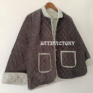 以下が含まれることがあります： ダイヤモンド模様のダークブラウンのキルティングジャケット。フロントポケットが2つ、ストライプのトリムが付いています。白い壁に木製のハンガーに掛けられています。「SATISFACTORY」の文字がジャケットにプリントされています。