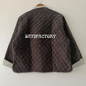 以下が含まれることがあります： ダイヤモンド模様のダークグレーのキルティングジャケット。 ジャケットにはストライプの襟と袖口が付いています。 「SATISFACTORY」の文字がジャケットの背面に白でプリントされています。 ジャケットは木製のハンガーに吊るされています。