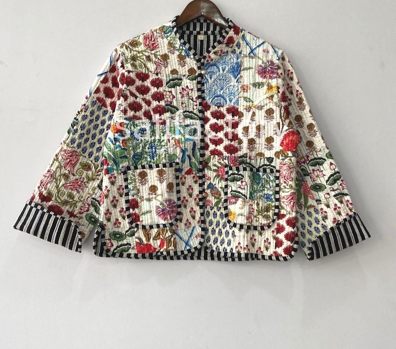 K&ouml;nnte beinhalten: Gesteppte Jacke mit floralem Patchwork-Design. Die Jacke zeigt eine Vielzahl farbenfroher Blumenmuster, darunter rote, blaue und rosa Blumen, auf wei&szlig;em Hintergrund. Die &Auml;rmel und der Saum sind mit schwarz-wei&szlig;en Streifen versehen.