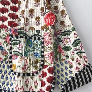 K&ouml;nnte beinhalten: Gesteppte Jacke mit Patchwork-Design mit Blumen- und geometrischen Mustern in Rot, Blau und Gelb auf wei&szlig;em Hintergrund. Die Jacke hat eine schwarz-wei&szlig; gestreifte Bord&uuml;re und eine Tasche.