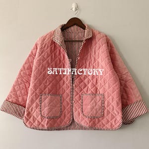 May include: A quilted pink jacket with a diamond pattern, two front pockets, and a striped trim. The jacket has a collared neckline and is buttoned up. The text "SKITIEFACTORY" is printed on the front.