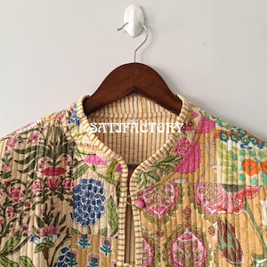 K&ouml;nnte beinhalten: Gesteppte Jacke mit Patchwork-Design mit Blumen- und Paisley-Mustern in Rosa, Blau, Gr&uuml;n und Gelb. Die Jacke hat einen Stehkragen und rosa Kn&ouml;pfe. Der Text "SATISFACTORY" ist auf dem Kragen sichtbar. Die Jacke h&auml;ngt an einem Holzb&uuml;gel.