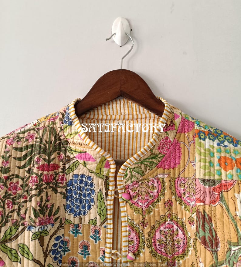 K&ouml;nnte beinhalten: Gesteppte Jacke mit Patchwork-Design mit Blumen- und Paisley-Mustern in Rosa, Blau, Gr&uuml;n und Gelb. Die Jacke hat einen Stehkragen und rosa Kn&ouml;pfe. Der Text "SATISFACTORY" ist auf dem Kragen sichtbar. Die Jacke h&auml;ngt an einem Holzb&uuml;gel.