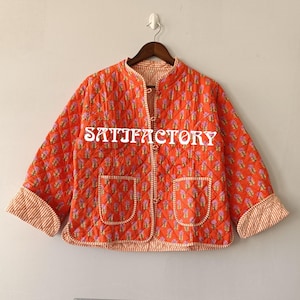 Peut inclure: Veste matelassée orange avec un motif floral et le mot "SATISFACTORY" en blanc. La veste a deux poches et des poignets et bordures rayés. Elle est suspendue à un cintre en bois.