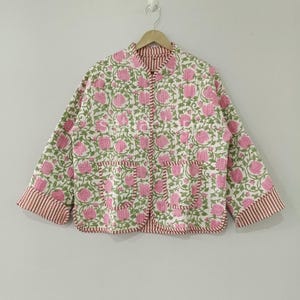 Op de afbeelding: Een gewatteerde jas met een bloemenpatroon in roze en groen op een witte achtergrond. De jas heeft een ronde kraag, twee voorzakken en gestreepte boorden aan de manchetten en zoom. De jas hangt aan een houten hanger.