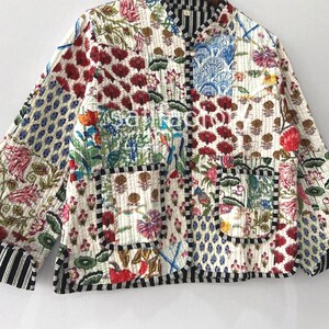 K&ouml;nnte beinhalten: Gesteppte Jacke mit Patchwork-Design mit floralen und geometrischen Mustern in Rot, Blau, Gr&uuml;n und Wei&szlig;. Die Jacke hat eine schwarz-wei&szlig; gestreifte Bord&uuml;re und einen Stehkragen. Die Jacke h&auml;ngt an einem Holzkleiderb&uuml;gel.
