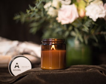 May include: A brown glass candle jar with a lit candle inside. The candle is surrounded by a bouquet of pink and white flowers.