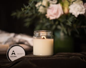 May include: A lit candle in a clear glass jar with a white wax interior. The candle sits on a dark, textured surface, with a partially visible lid nearby. The lid has the word "ADORN" and a logo on it. A bouquet of flowers is in the background.