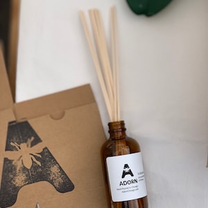 May include: A brown glass bottle with a white label that says "Adorn" and features a bee illustration. The label also says "Volcano Ash Diffuser" and "Hand Poured in Chicago". The bottle is sitting on a brown cardboard box with a black bee illustration. There are wooden sticks in the bottle.