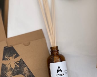 May include: A brown glass bottle with a white label that says "Adorn" and features a bee illustration. The label also says "Volcano Ash Diffuser" and "Hand Poured in Chicago". The bottle is sitting on a brown cardboard box with a black bee illustration. There are wooden sticks in the bottle.