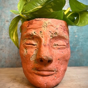 May include: A terracotta-colored face planter with closed eyes, a nose, and a smiling mouth. The planter is filled with green leafy plants. The planter has a rough, textured surface and sits on a wooden surface.