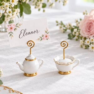 May include: Two miniature white ceramic place card holders with gold trim, one shaped like a teapot and one like a teacup, each featuring a gold spiral wire clip. A white rectangular place card with floral accents and the name Eleanor is displayed.