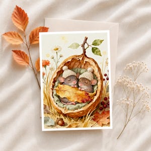 May include: A greeting card featuring a watercolor illustration of two small mice wearing cream-colored knit hats, sleeping inside a large walnut shell lined with green fabric and covered by a golden autumn leaf, surrounded by wildflowers and berries.