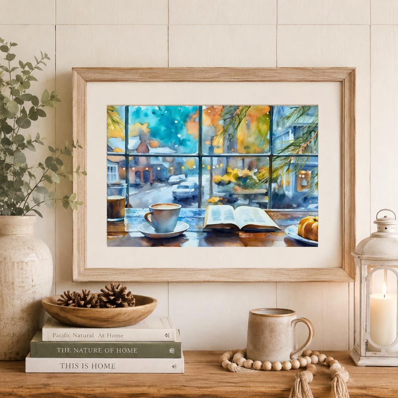 May include: A framed watercolor print depicting a window view of a snowy street with a coffee cup, an open book, and a pastry on a wooden table. The artwork is displayed in a light wood frame, surrounded by a vase, pinecones, and a lantern.