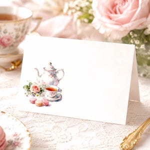 May include: A white folded greeting card featuring a watercolor illustration of a floral teapot, a matching teacup filled with tea, and three small pink and beige macarons arranged on the lower left side of the card's front surface.