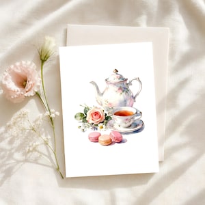 May include: A greeting card featuring a watercolor illustration of a floral teapot, a matching teacup filled with tea on a saucer, a single pink rose, and three colorful macarons. The artwork is centered on a white card with a plain envelope.
