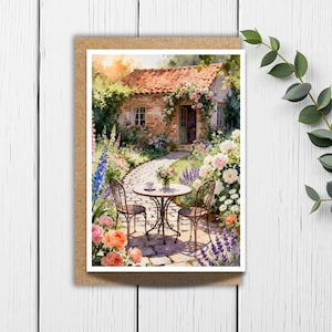 May include: A watercolor greeting card featuring a charming cottage scene. The image depicts a stone path leading to a brick cottage adorned with flowers. A small table and chairs are set in a garden filled with colorful blooms. The card is set against a white wooden background.