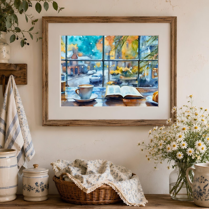 May include: A framed watercolor print depicting a cozy indoor scene with an open book and a cup of coffee on a table, overlooking a blurred street view through a window. The artwork features vibrant blue, yellow, and brown tones in a landscape orientation.