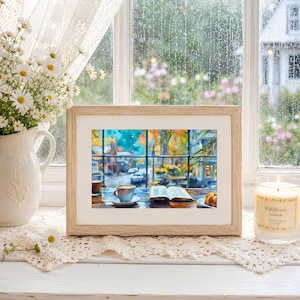 May include: A framed watercolor print depicting a cozy indoor scene with a coffee cup and an open book on a table, set against a rainy window view. The artwork is displayed in a light wood frame, accompanied by a white pitcher of daisies and a candle.