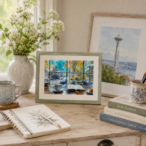 May include: A framed watercolor print depicting a cozy window view with a coffee cup and an open book. The artwork features vibrant blue and yellow tones, set within a light green wooden frame, displayed on a wooden surface with books and a vase.