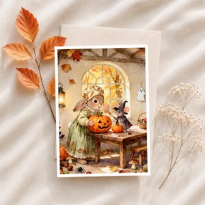 May include: A greeting card illustration featuring a rabbit in a floral bonnet and green dress holding a carved jack-o'-lantern, and a mouse in a witch hat and cape standing at a wooden table with a small pumpkin, set in a room with autumn leaves.