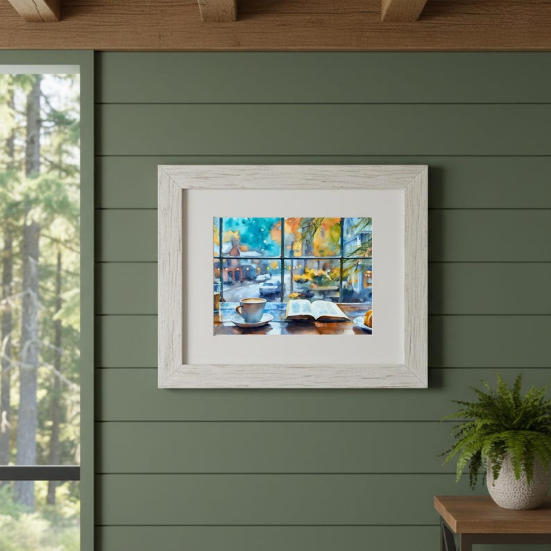 May include: A framed watercolor print featuring a view through a window of a street scene with a coffee cup and an open book on a table in the foreground. The artwork is encased in a white wooden frame with a white mat board border.
