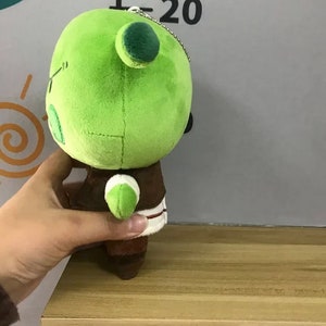 Peut inclure: Une peluche verte avec une t&ecirc;te ronde, deux petites oreilles et une tenue marron. Le jouet est tenu par une main et est pos&eacute; sur une &eacute;tag&egrave;re en bois. L'arri&egrave;re-plan est un mur gris clair avec une &eacute;chelle de mesure.