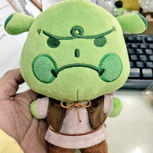 Peut inclure: Une peluche verte avec une expression grincheuse, avec des d&eacute;tails brod&eacute;s. Elle a de petites oreilles, un gilet marron et des chaussures marron. Le jouet est tenu dans la main d'une personne.