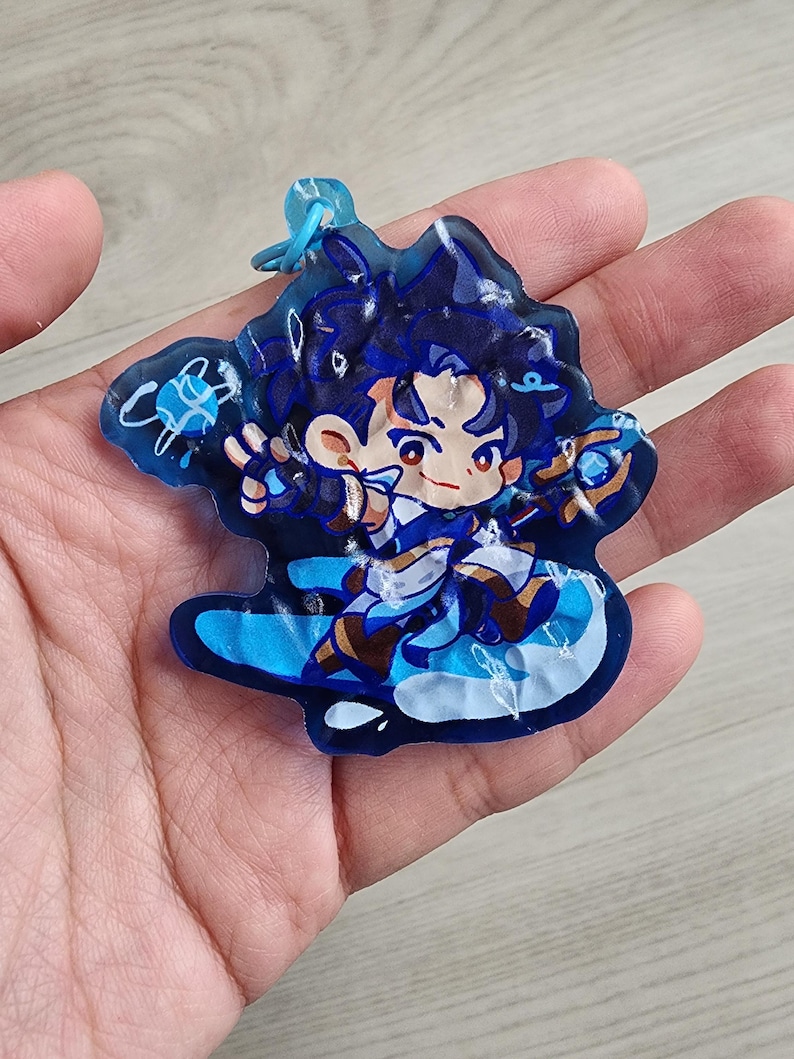 May include: A translucent blue acrylic keyring featuring a stylised anime character with dark hair, wearing a white and blue outfit, holding a staff, and surrounded by swirling blue energy effects, attached to a small blue plastic ring connector.
