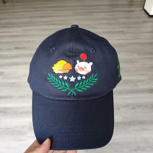 Puede incluir: Gorra de béisbol azul marino con personajes de dibujos animados bordados y un diseño de corona de laurel verde. La gorra tiene una visera curva y una corona estructurada.