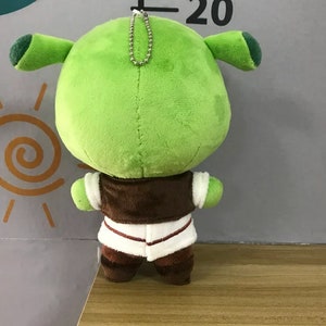 Peut inclure: Une peluche verte repr&eacute;sentant un ogre, avec de grandes oreilles et un corps rond. L'ogre porte un gilet marron sur une chemise blanche et un pantalon marron. Une cha&icirc;ne argent&eacute;e est attach&eacute;e au sommet du jouet.