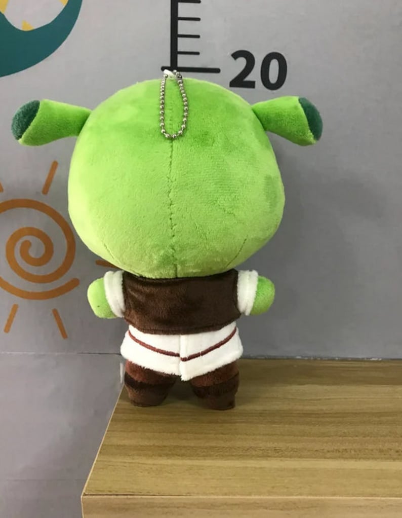 Peut inclure: Une peluche verte repr&eacute;sentant un ogre, avec de grandes oreilles et un corps rond. L'ogre porte un gilet marron sur une chemise blanche et un pantalon marron. Une cha&icirc;ne argent&eacute;e est attach&eacute;e au sommet du jouet.