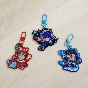 May include: Three acrylic keychains featuring anime-style characters. The left keychain is red with a character and a small blue creature. The centre keychain is dark blue with a space-themed character. The right keychain is light blue with a character.