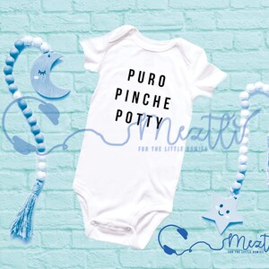 May include: A white baby grow with black text that reads "PURO PINCHE POTTY". The grow is on a blue brick background with blue and white beaded garlands and a blue wooden star and moon.