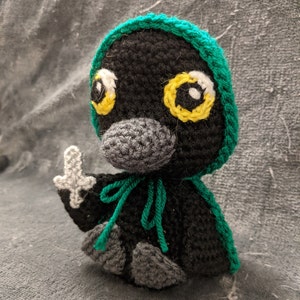 May include: A crocheted black and green bird wearing a green hooded cloak. The bird has large yellow eyes and a gray beak. The bird is holding a white object in its right hand.