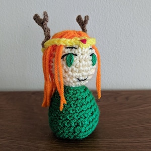 May include: A crocheted doll with orange hair, brown antlers, and a yellow and red crown. The doll is wearing a green dress and has green eyes.