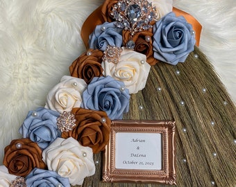 Peut inclure: Un arrangement floral brun, blanc et bleu avec des accents de perles orne un balai traditionnel. Un petit cadre doré avec le texte "Adrian & Dalena October 21, 2023" est fixé au balai.