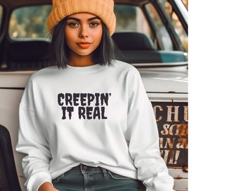 May include: White sweatshirt with the text "CREEPIN' IT REAL" in a dark, spooky font. The sweatshirt is worn by a person wearing a yellow beanie and blue jeans.