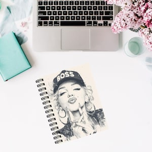 May include: Spiral-bound notebook featuring a black and white illustration of a woman wearing a "BOSS" baseball cap. The notebook is open, displaying the artwork. A laptop, a teal book, and pink flowers are also visible.