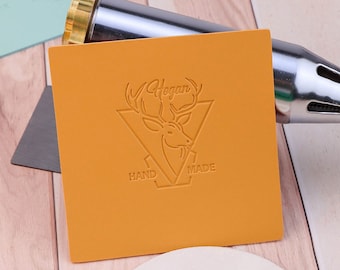 Könnte beinhalten: Eine Nahaufnahme eines Lederstempels mit einem Hirschkopfdesign und dem Text "Hogan Hand Made". Der Stempel befindet sich auf einer gelben Lederoberfläche.