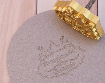 Könnte beinhalten: Ein goldfarbener Metallstempel mit einem Baumdesign und den Worten "Handmade with Love" in einer dekorativen Schriftart. Der Stempel wird verwendet, um einen Abdruck auf einem Stück hellbraunem Leder zu erstellen. Der Abdruck lautet "Handmade with Love" und "Greetings".