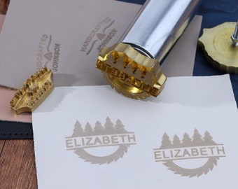 Könnte beinhalten: Zwei goldfarbene Metallstempel mit einem Design aus einer Reihe von Kiefern und dem Wort "ELIZABETH" in einem Banner. Die Stempel befinden sich auf einer weißen Oberfläche.